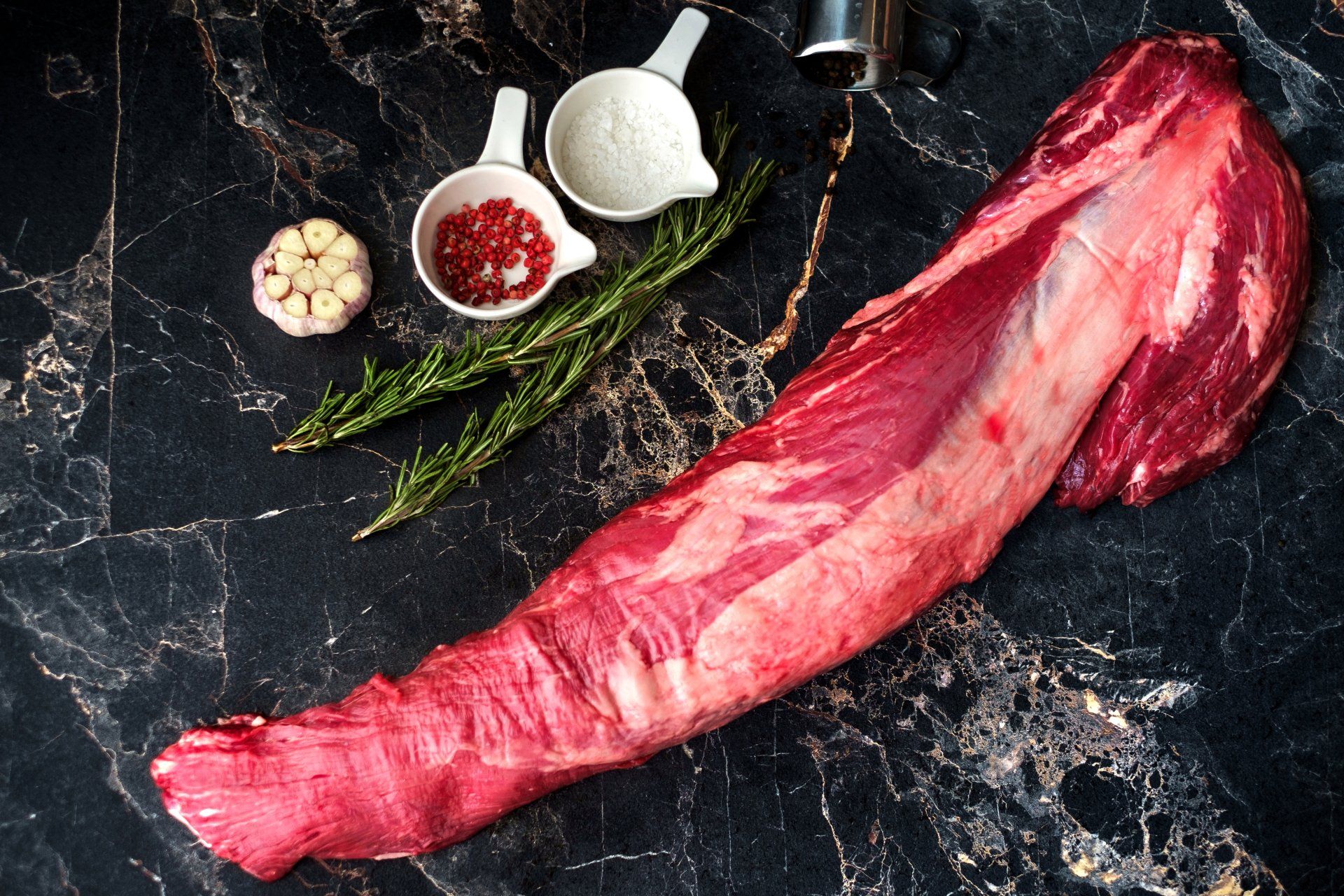 A large piece of raw beef is sitting on a marble surface next to a hammer.