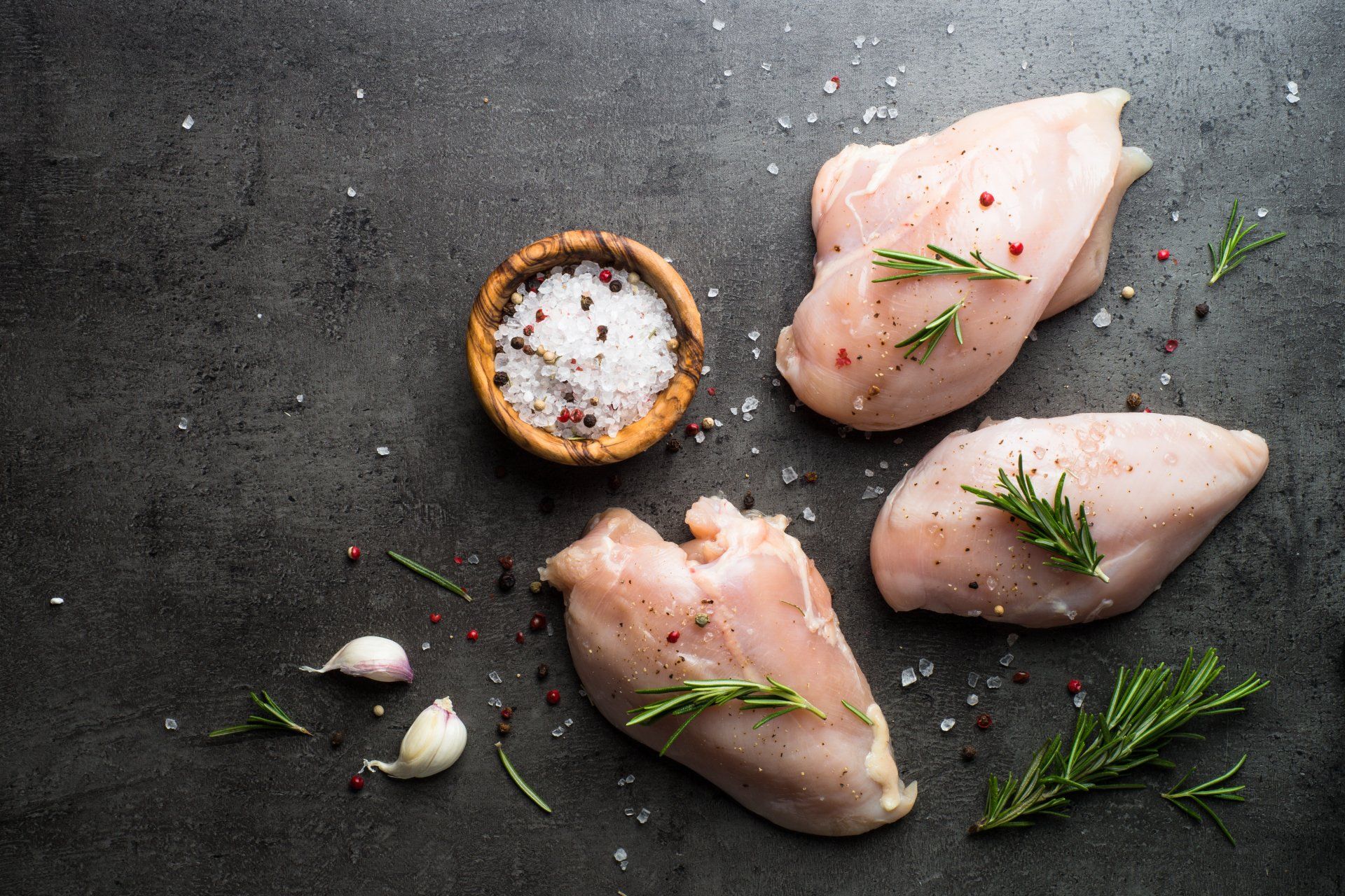 Raw chicken breasts with spices and herbs on a black table.