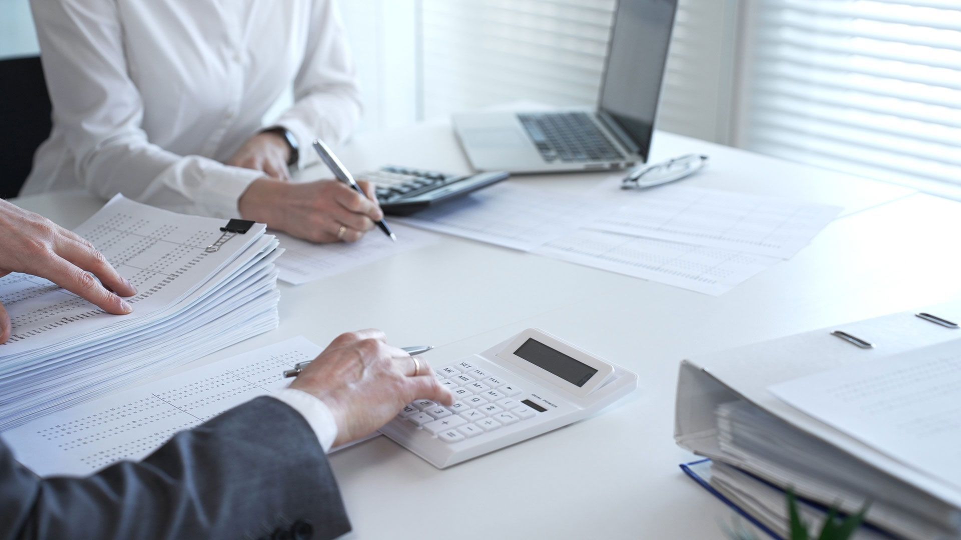 People reviewing documents at a bright office table with laptops, calculator, and stacked papers