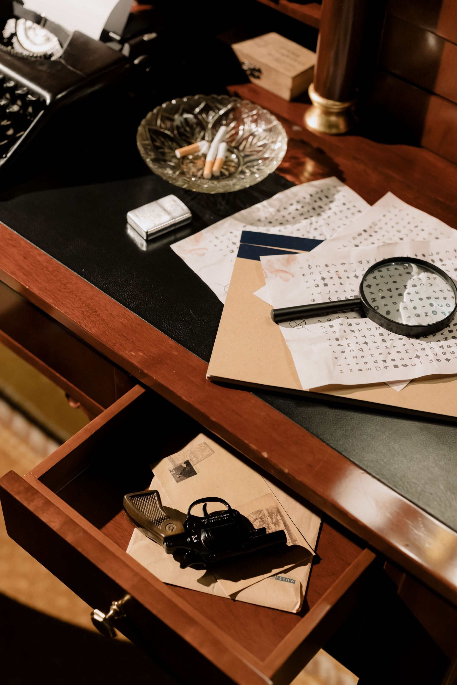 Desk with scattered papers, magnifying glass, ashtray, and a drawer containing keys and a black object.