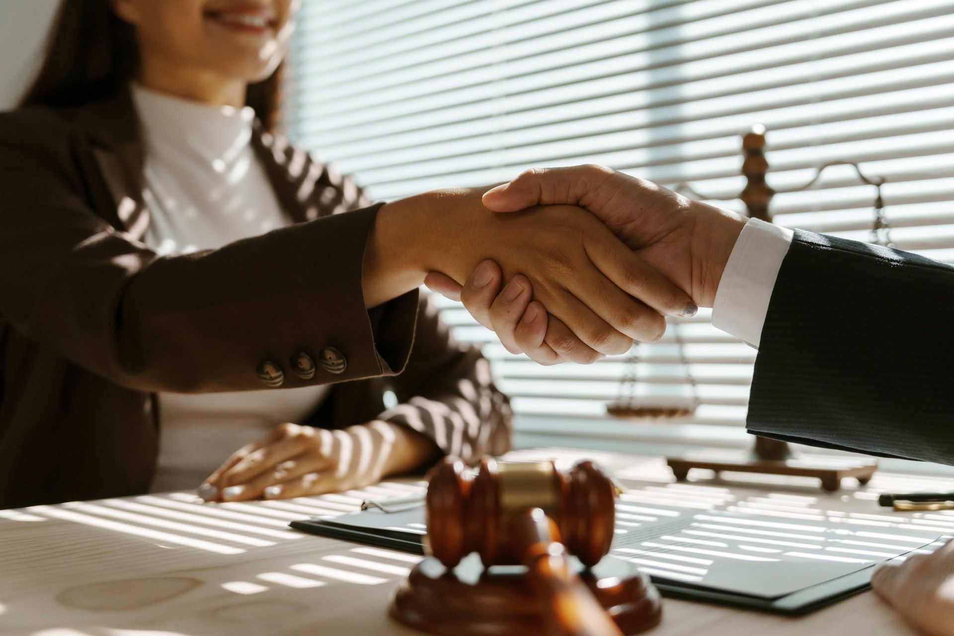 Two people shake hands across a desk in a bright office with a gavel in the foreground.