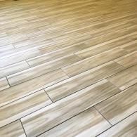 Light-colored wood-look ceramic floor tiles with a linear grain pattern, installed in a staggered horizontal layout.