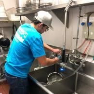 A person in a blue t-shirt and grey cap washing metal cookware in a large industrial commercial kitchen sink.