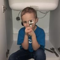 A child in a blue shirt sits under a sink, holding an adjustable wrench up to their eye like a monocle.