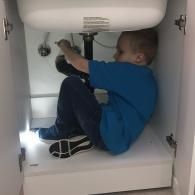 A person in a blue shirt and jeans sits inside a dark cabinet under a sink, working on the plumbing pipes.