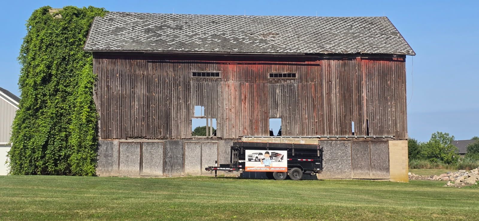 Old weathered barn with ivy on one side and a parked trailer in front, under a clear sky.