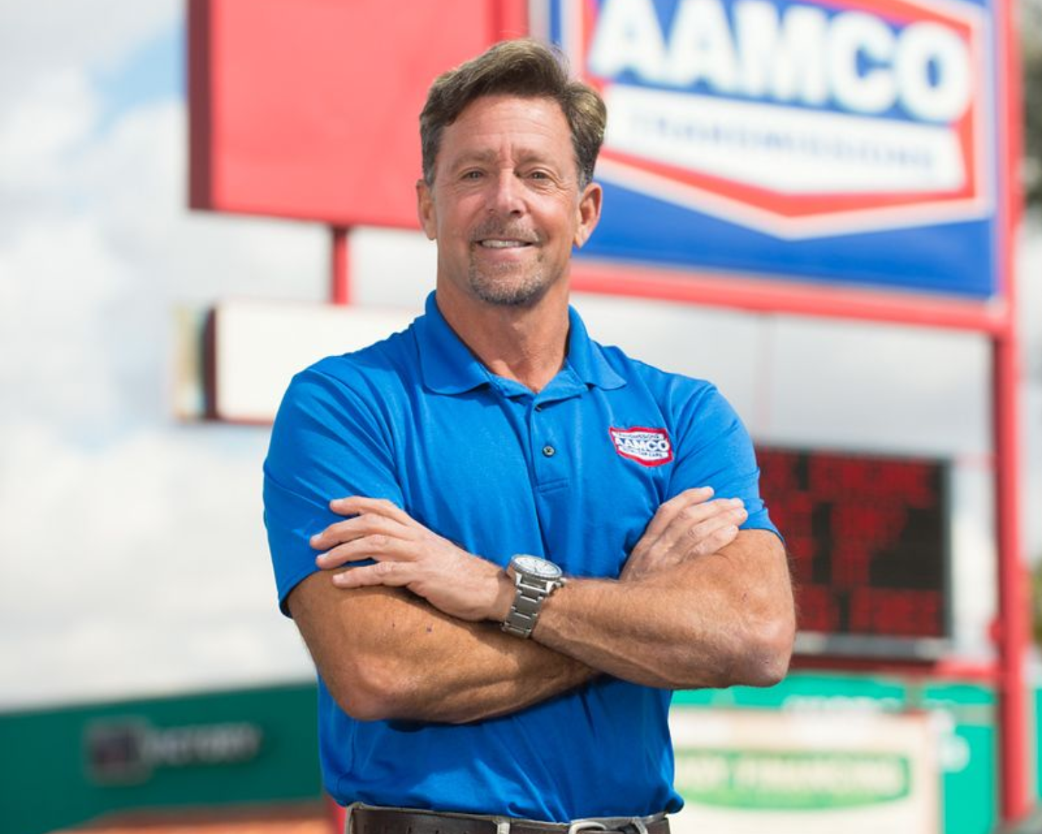 A person in a blue polo shirt with arms crossed, smiling in front of an AAMCO signage outdoors.