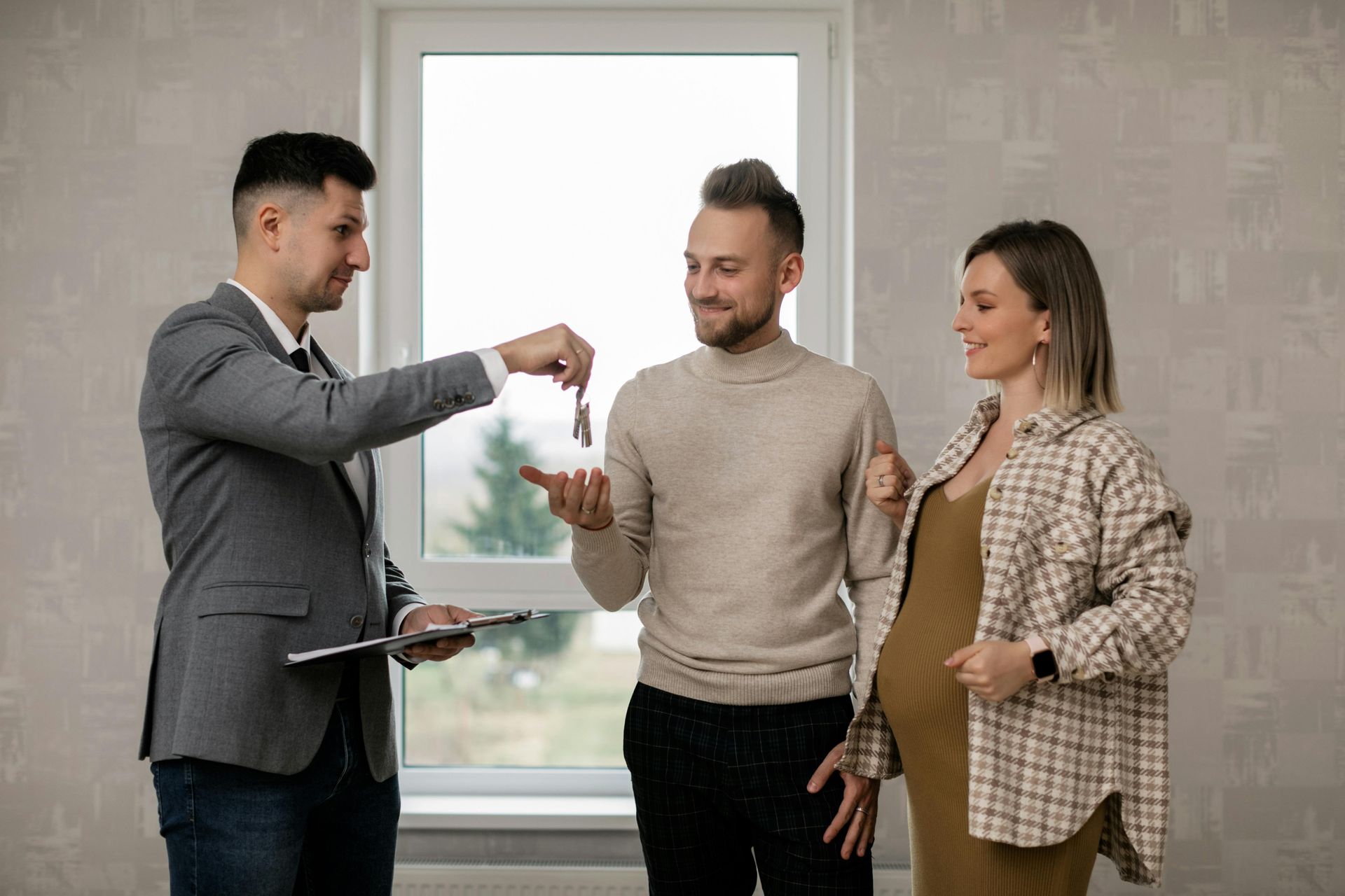 Real estate agent hands keys to a smiling couple, one pregnant, in a new home.