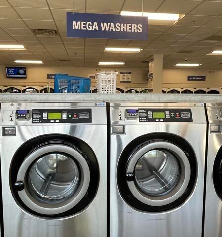 Row of stainless steel mega washers in a laundromat with a blue “MEGA WASHERS” sign overhead