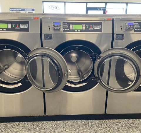 Row of stainless steel front-loading washing machines with open doors in a laundromat