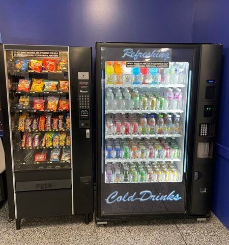 Two vending machines against a blue wall, one snacks and one cold drinks, lit in a hallway.