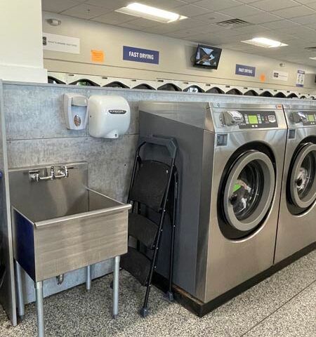 Laundromat with washing machines, stainless sink, hand dryer, and folded chairs against a tiled wall.