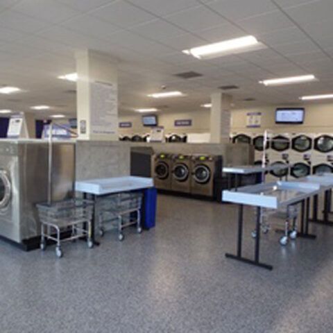 Laundry room with rows of washers and dryers, folding tables, and utility carts under bright fluorescent lights