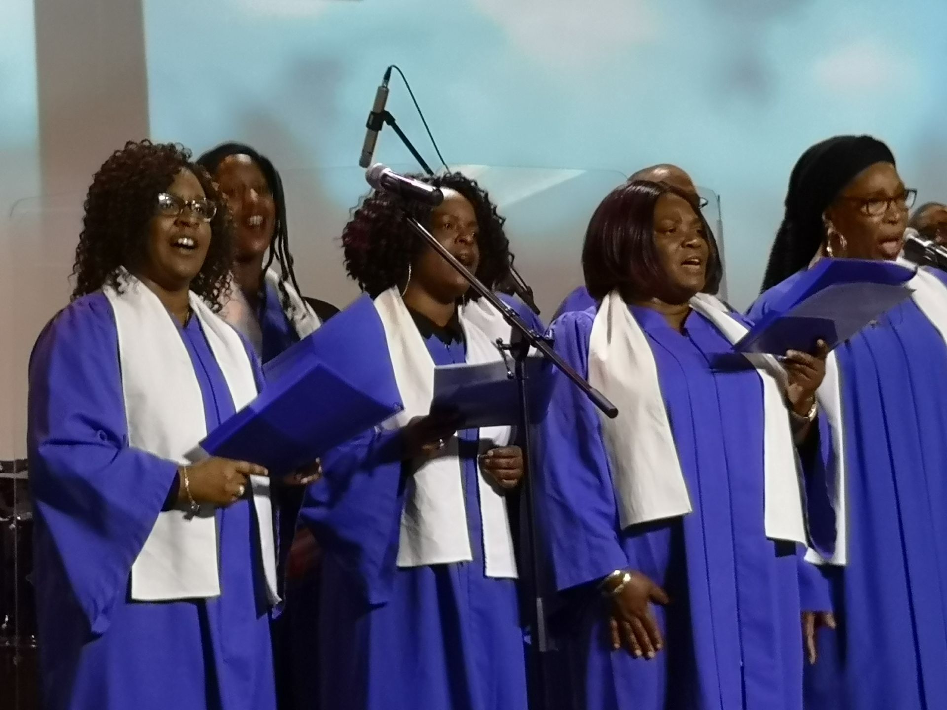 Choir in blue robes with white stoles sing into microphones.