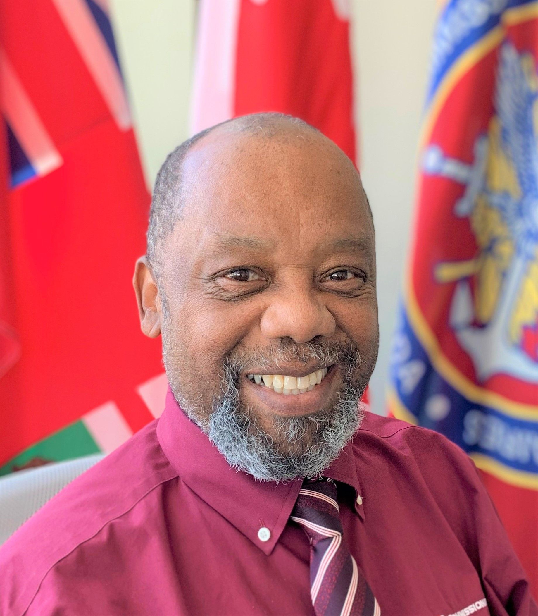 Man with a gray beard smiles, wearing a burgundy shirt and tie, with flags in the background.