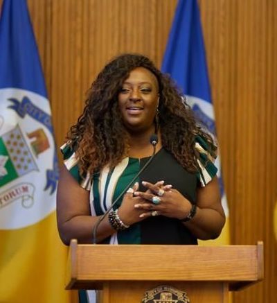 Woman speaking at a podium, wearing a dress, with state flags in the background.
