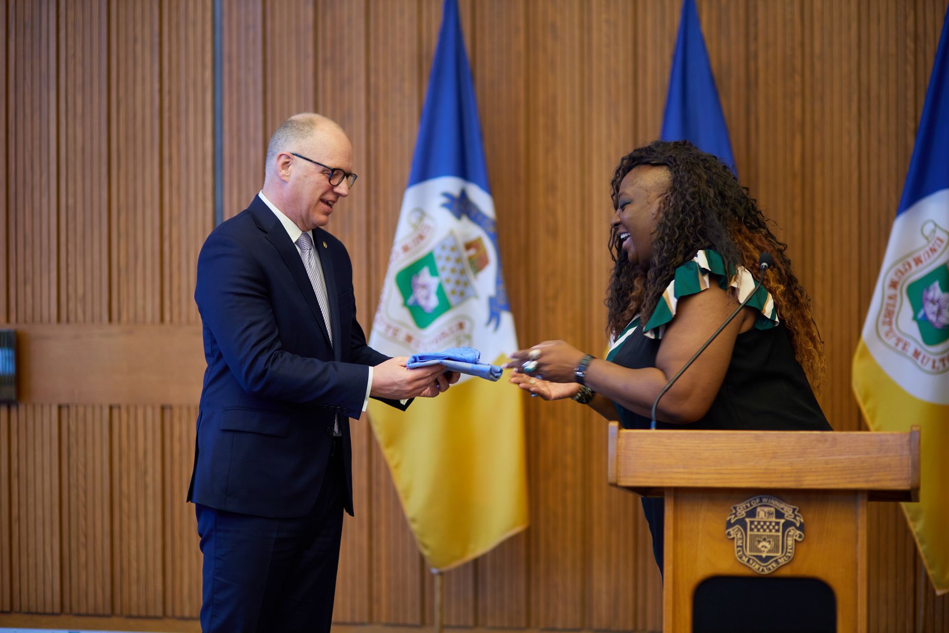 Man in suit hands a document to a woman at a podium; flags in background.