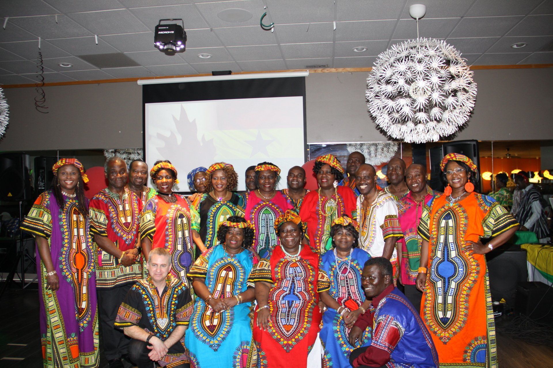 Group of people in colorful patterned clothing posing indoors. Bright lights and a projector screen are in the background.