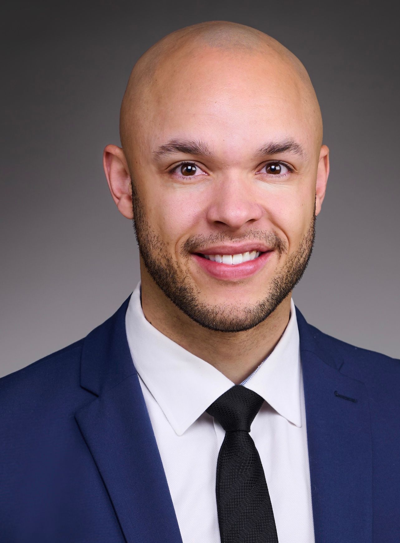 Man in a blue suit smiles at the camera against a gray background.
