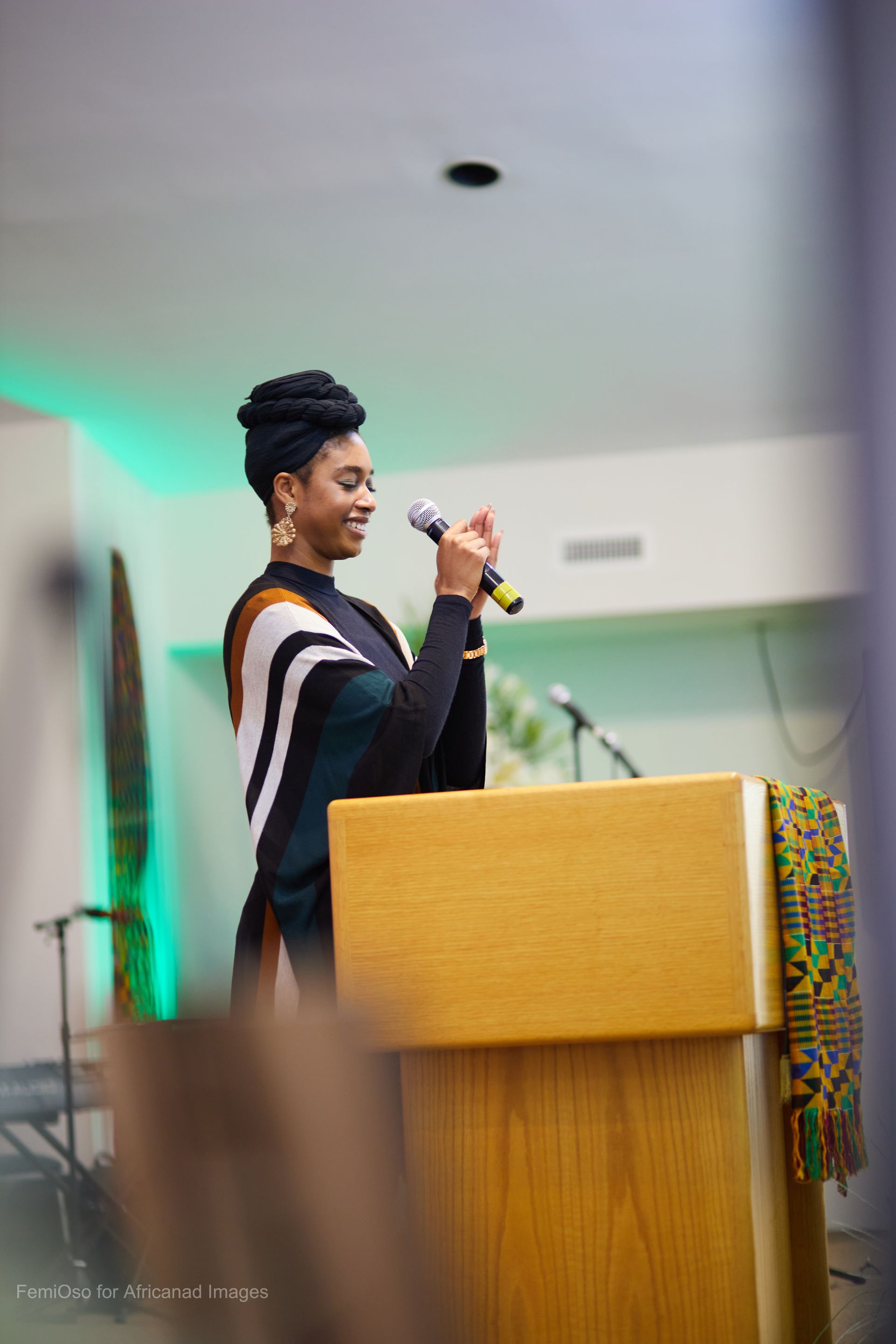 Woman speaking at a podium, smiling, holding a microphone. Green and white lighting in the background.