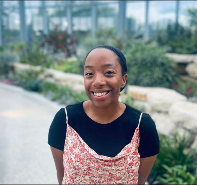 Woman smiles in front of plants, wearing a black top and floral dress.