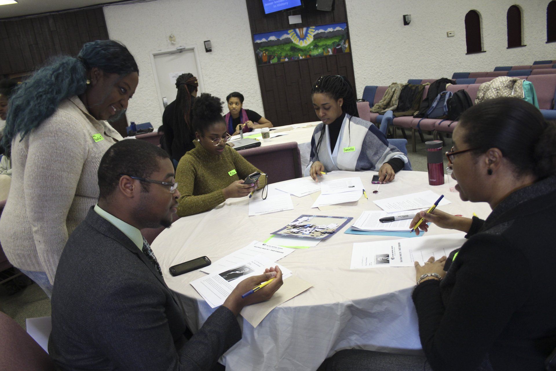 People seated at round tables collaborating, writing, and looking at papers in a meeting room.