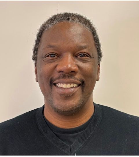 Man with short, graying hair smiles, wearing a black shirt, against a light wall.