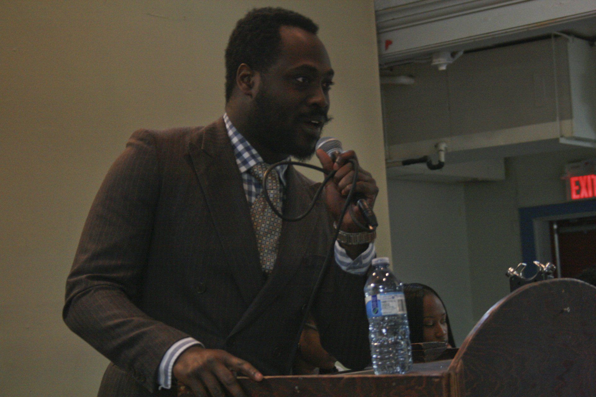 Man in brown suit speaks into a microphone at a podium. Water bottle sits nearby.