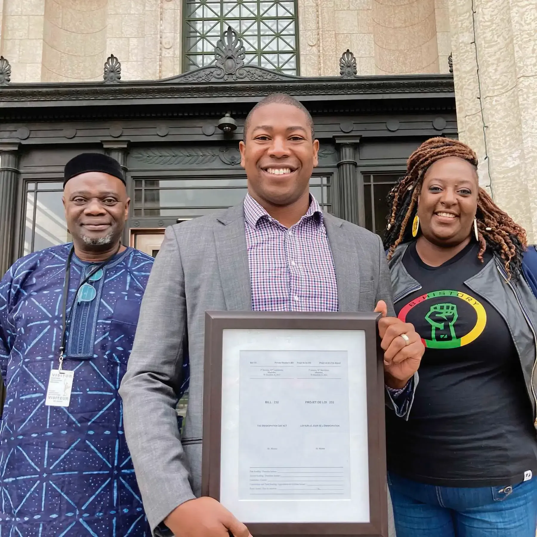 Man holding framed document flanked by two people, smiling in front of a building.