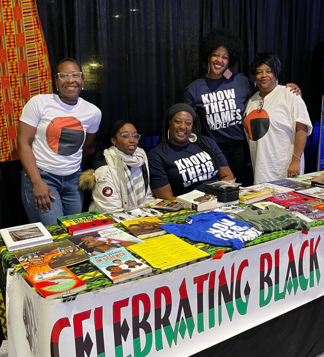People at a table displaying books and merchandise, with a banner that says,