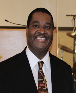Man in suit smiles, standing near a podium, with a microphone and drums in the background.