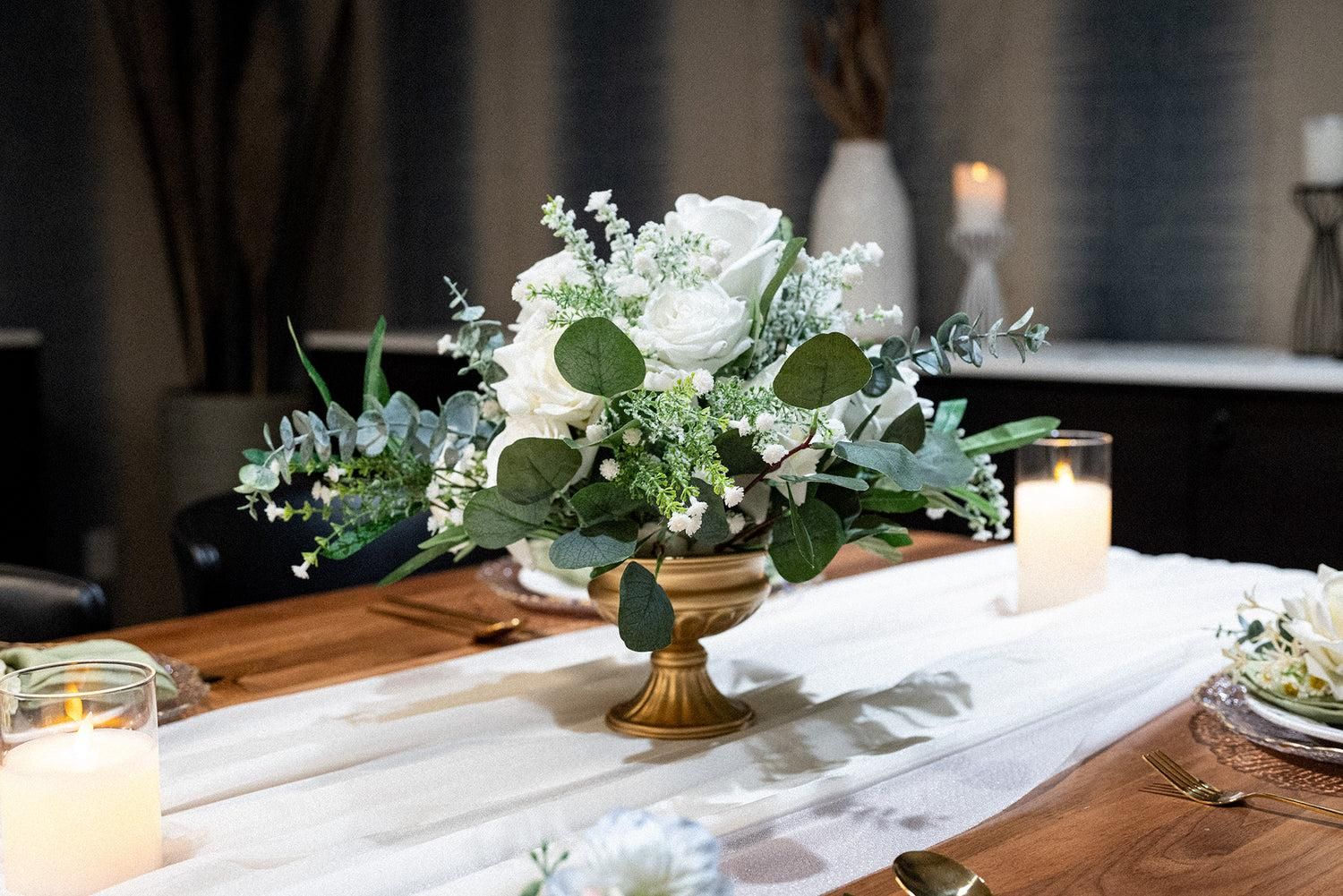 White floral centerpiece in a gold vase on a candlelit table with a runner and wooden accents