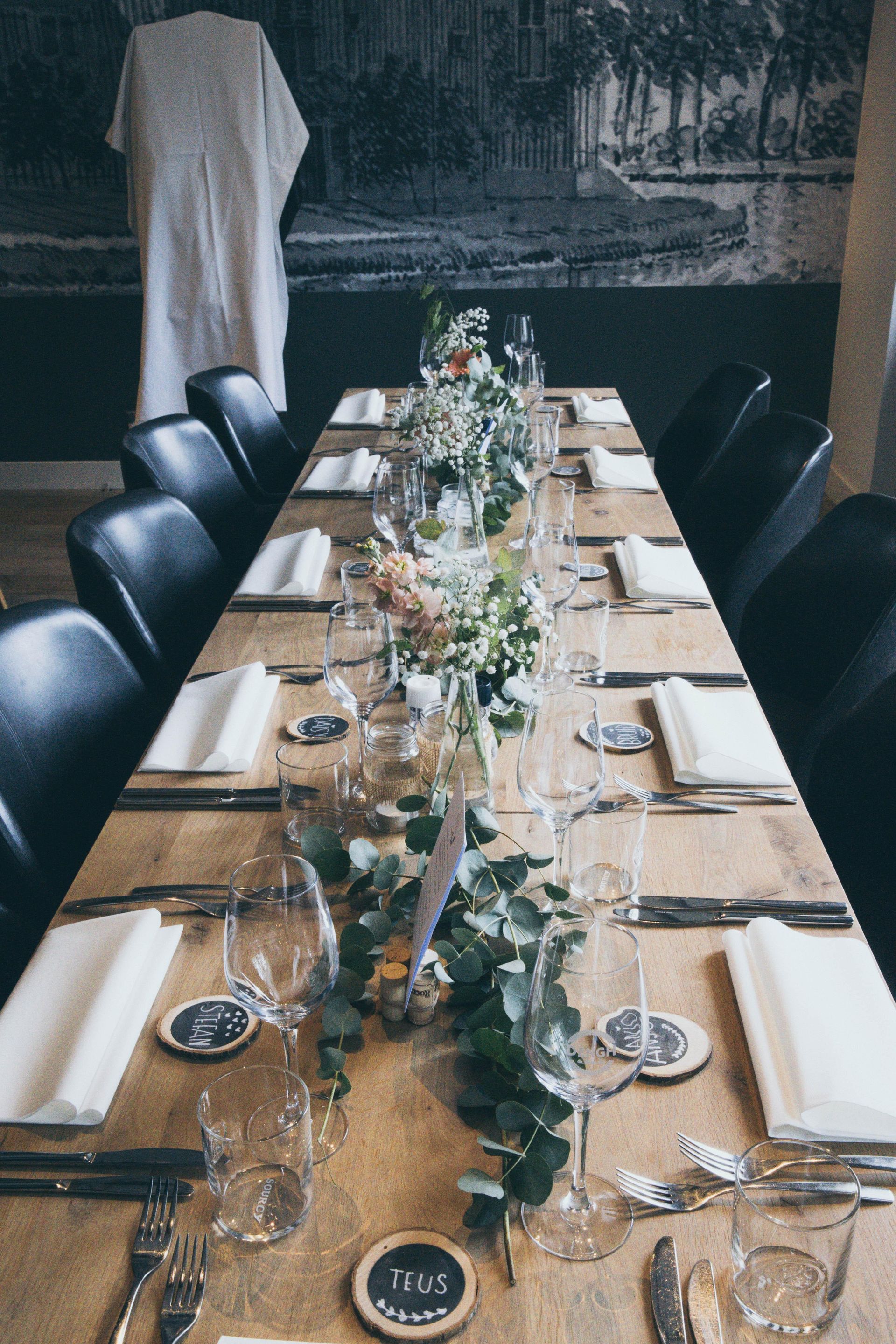 Long banquet table with black chairs, white place settings, and a floral greenery centerpiece in a rustic room