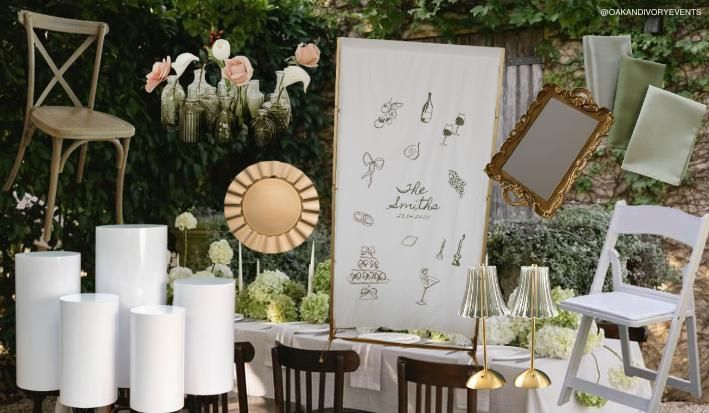 Wedding decor display with mirrors, candle pillars, white chairs, and floral arrangements against a garden backdrop