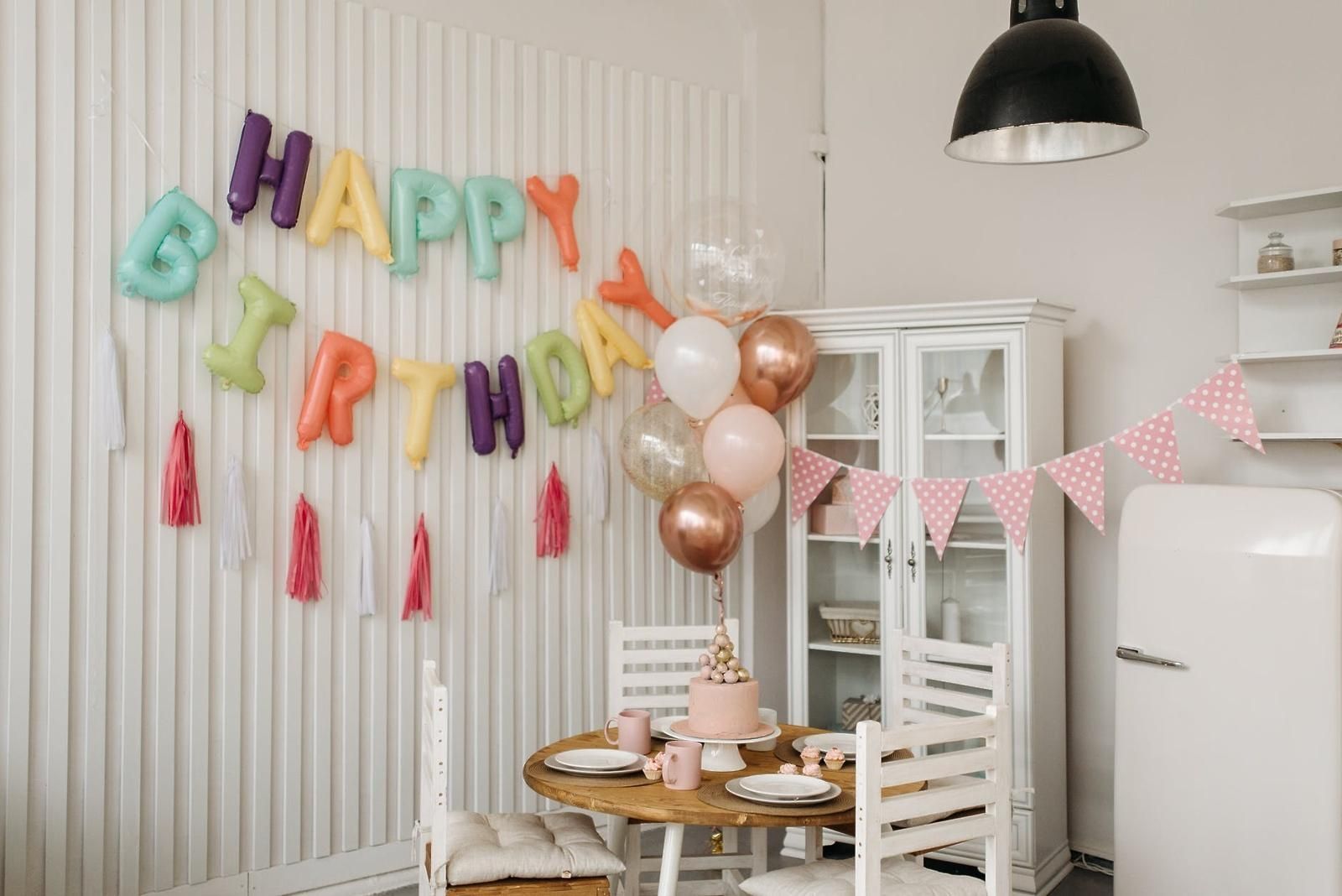 Pastel birthday party setup with balloon letters, rose-gold balloons, cake, and bunting in a white dining room