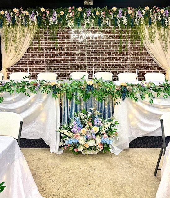 Flowered wedding sweetheart table with white drapes, greenery, pastel florals, and brick backdrop