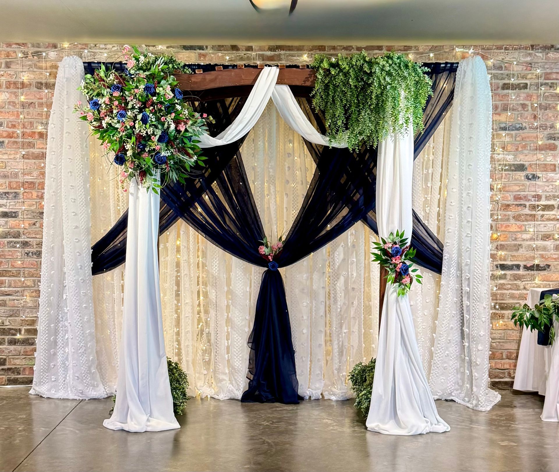 Decorated wedding arch with white and black drapes, floral greenery, and a brick backdrop