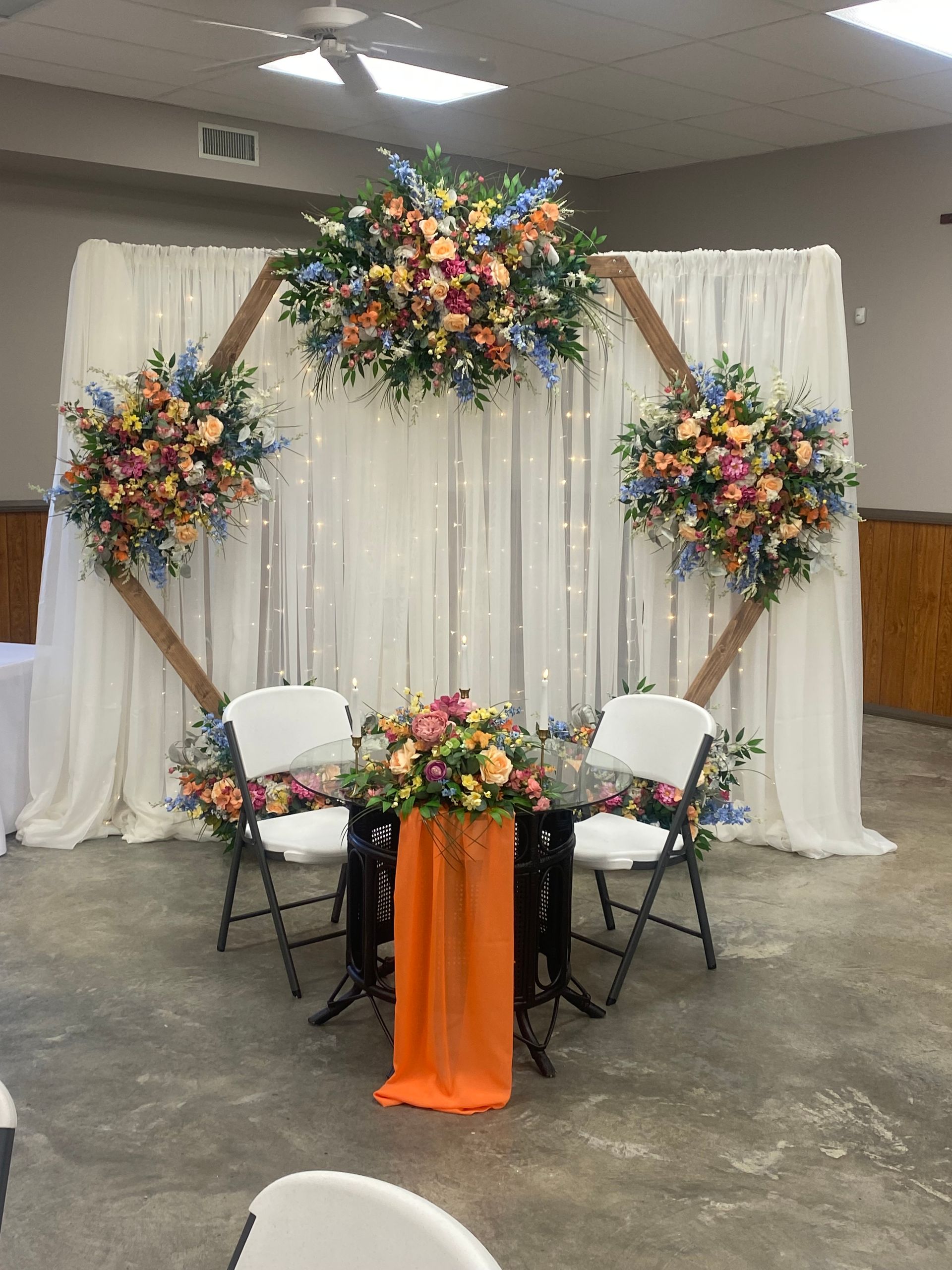 Decorated wedding backdrop with floral arches, draped white curtains, and two chairs around a small orange table