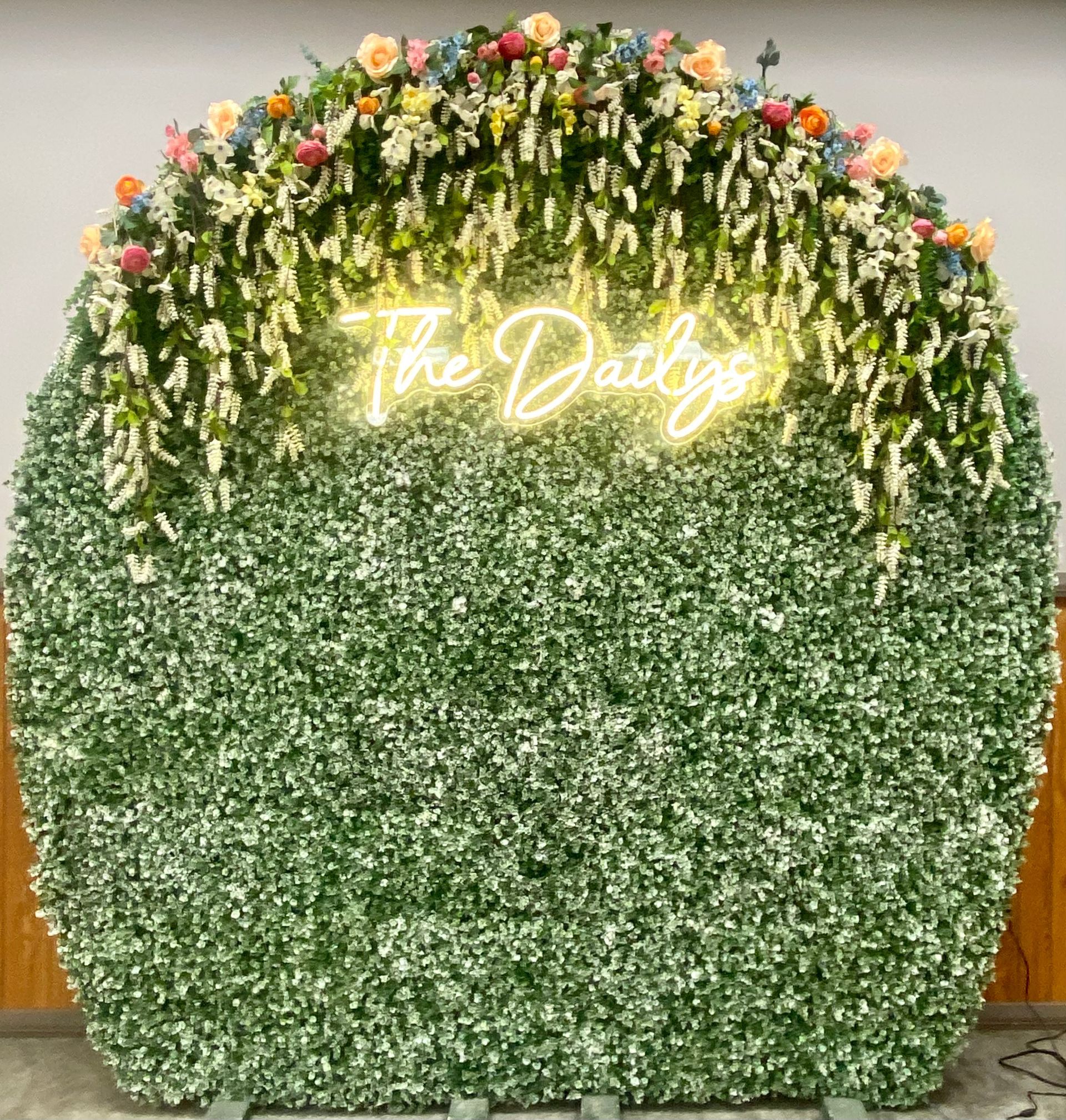 Green floral wall with white hanging flowers and a neon “The Daily” sign at top center