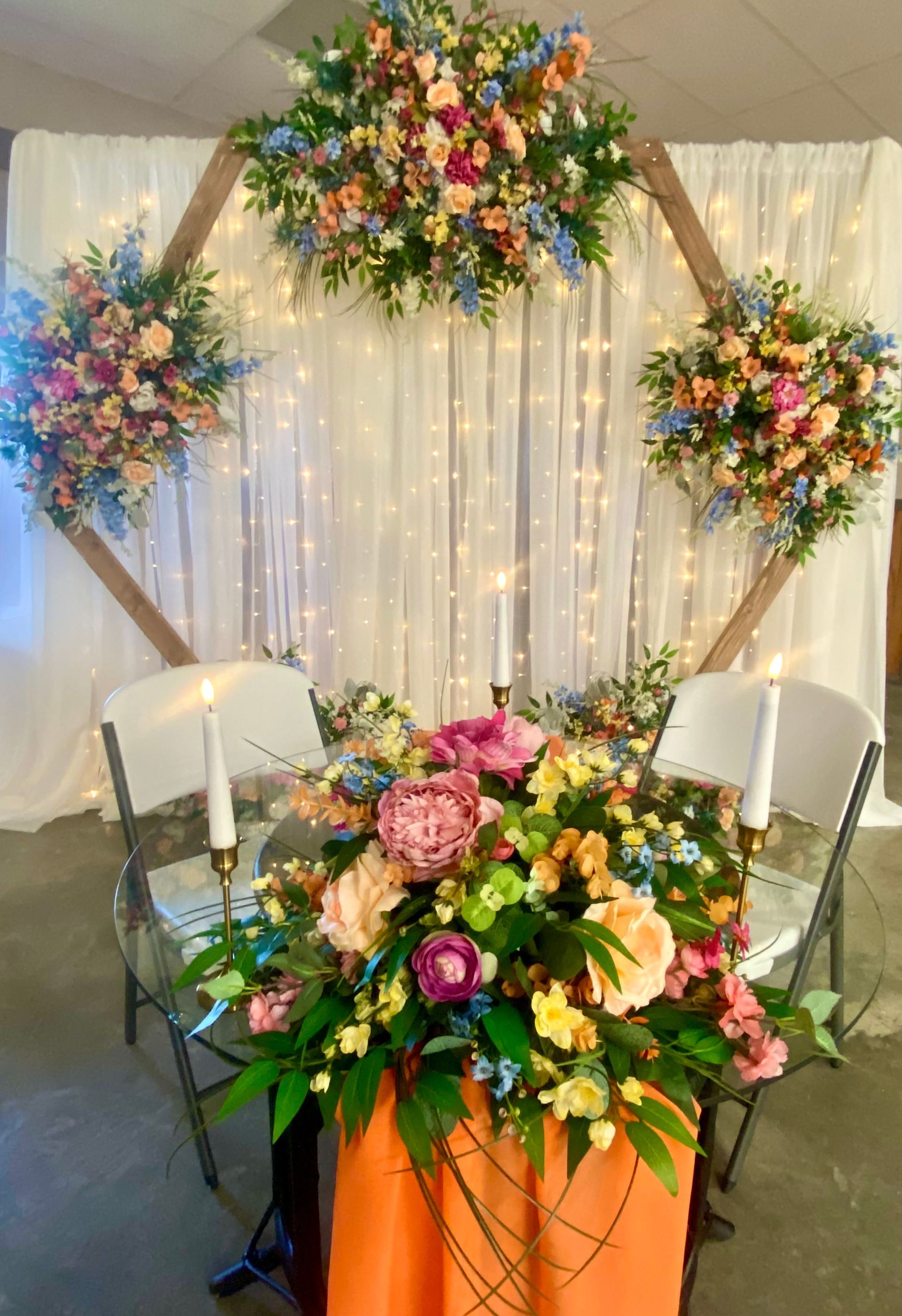 Floral wedding display with arch, bouquets, white chairs, and orange table centerpiece in a tented setting