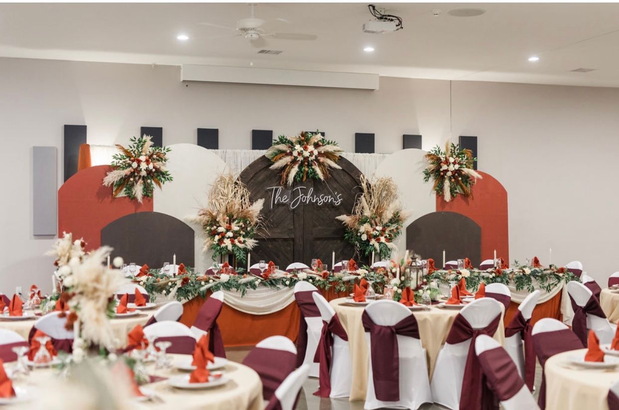 Elegant banquet hall set for a celebration with white and burgundy table decor and floral stage backdrop