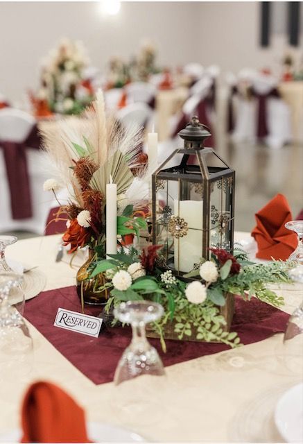 Rustic wedding table centerpiece with lantern, candles, greenery, and burgundy napkins on a banquet table