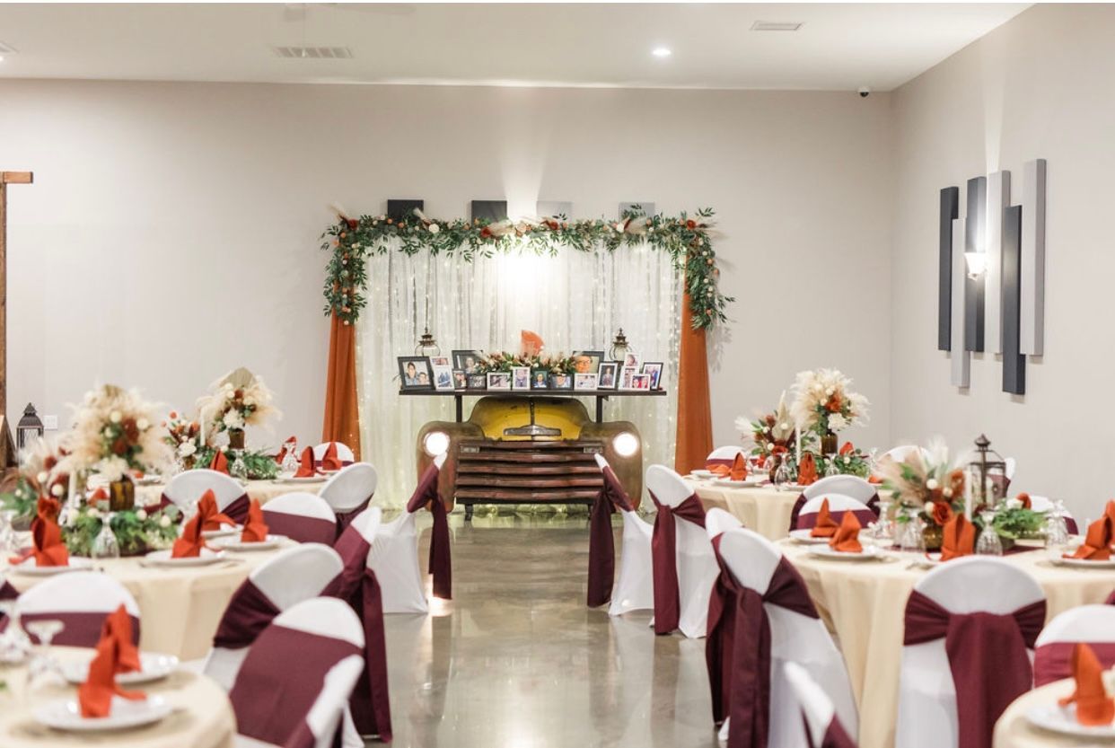 Decorated banquet hall with round tables, burgundy chair covers, and a floral arch backdrop
