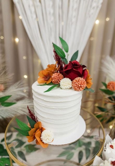 White tiered cake with orange and burgundy flowers on a mirrored table at a decorated event backdrop