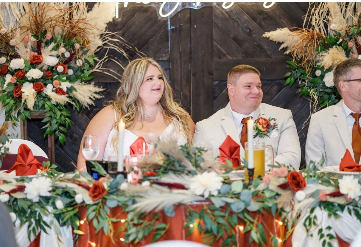 Three seated people at a decorated wedding reception table with floral arrangements and red napkins