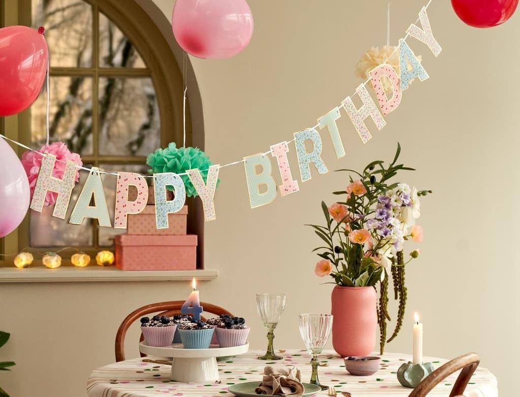 Pastel birthday table with balloon garland, “HAPPY BIRTHDAY” banner, cake, and flowers