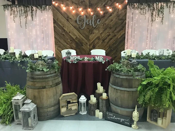 Rustic wedding backdrop with string lights, greenery, barrels, lanterns, and maroon table decor