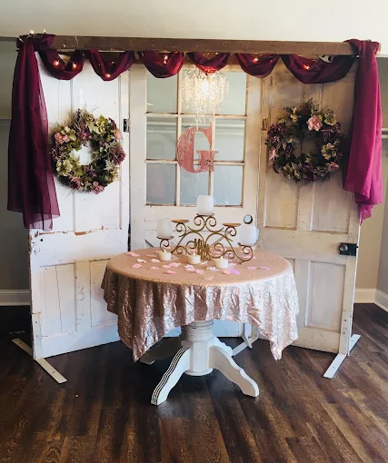 Decorated dessert table with pink and burgundy drapery, wreaths, and a monogrammed centerpiece on dark wood floor