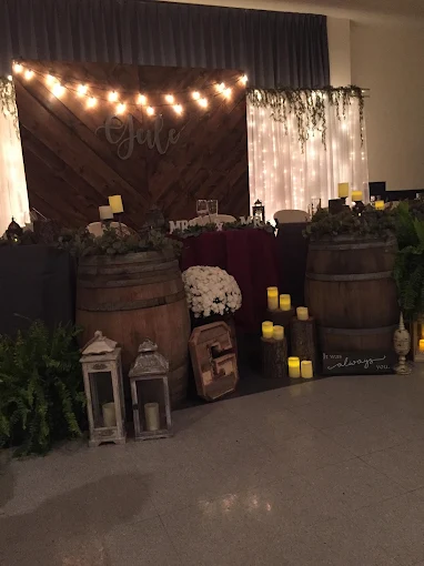 Rustic indoor event display with barrels, lanterns, flowers, candles, and string lights against dark drapes