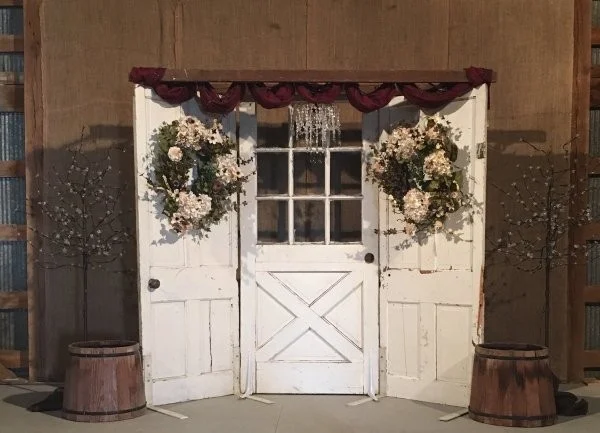 White rustic double doors with flower wreaths and barrels on either side under a burgundy awning